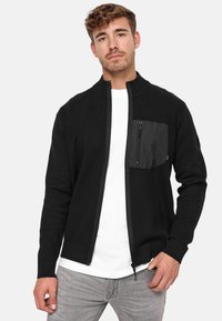 Sort lynlåset cardigan lavet af strikket stof med en struktureret forlommes og ribkantede ærmer. Båret over en hvid t-shirt, parret med lysegrå jeans.