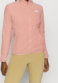 Fleecejack met zachte roze kleur, hoge kraag, volledige rits, zijzakken en wit logo-accent op de borst; gladde textuur.