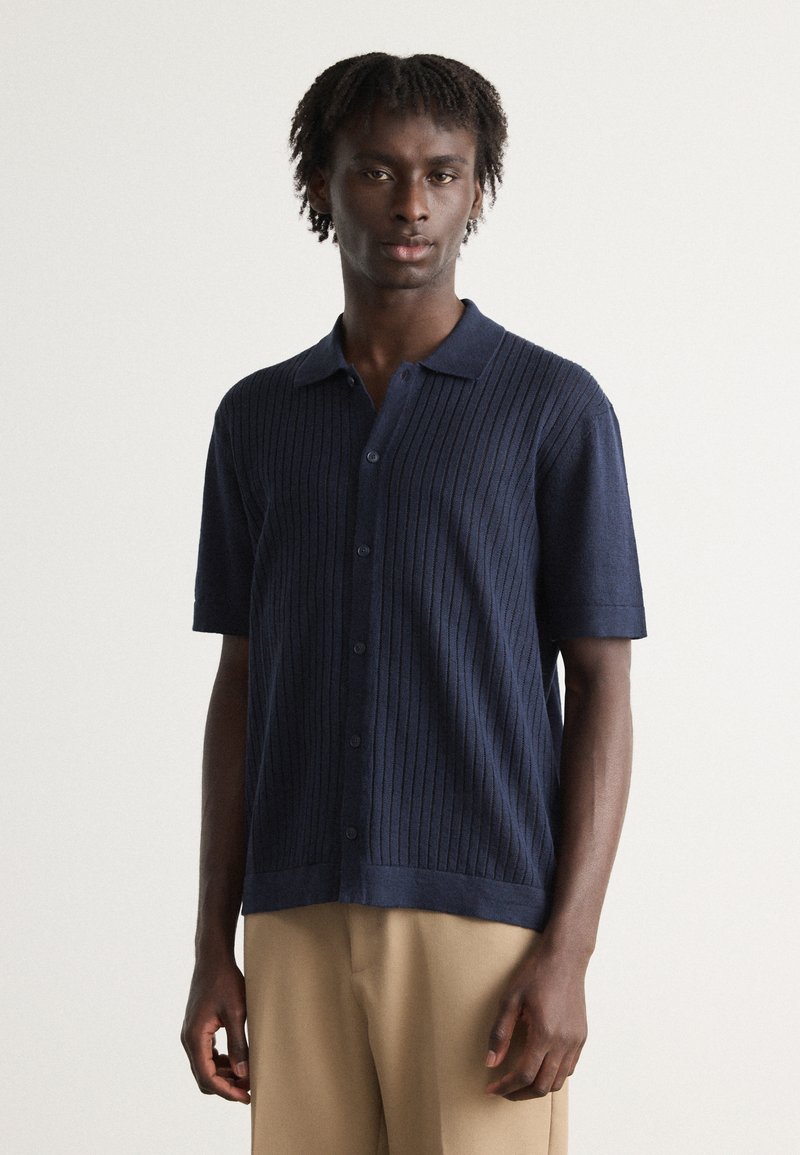 Polo shirt bleu marine a maniche corte con coste verticali, chiusura con bottoni e vestibilità comoda. Abbinato a pantaloni beige.