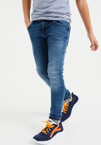 Slim-Fit blaue Jeans aus Denim mit leichter Ausbleichung, ausgestattet mit Fronttaschen und kontrastierenden Nähten. Kombiniert mit navyfarbenen Sneakers und orangen Schnürsenkeln.
