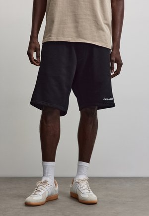 Mann trägt schwarze knielange Shorts, beiges T-Shirt, weiße Socken und weiße Sneakers mit Gummisohlen, steht auf einem grauen Boden.