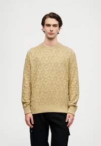 Μπεζ πλεκτό πουλόβερ με υφασμάτινο επαναλαμβανόμενο λογότυπο, στρογγυλή λαιμόκοψη και ριγέ μανίκια και στρίφωμα. Συνδυασμένο με μαύρο παντελόνι.