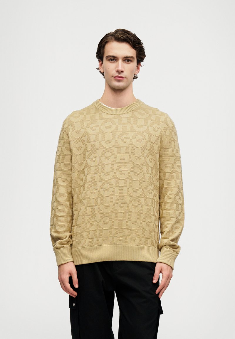 Μπεζ πλεκτό πουλόβερ με υφασμάτινο επαναλαμβανόμενο λογότυπο, στρογγυλή λαιμόκοψη και ριγέ μανίκια και στρίφωμα. Συνδυασμένο με μαύρο παντελόνι.