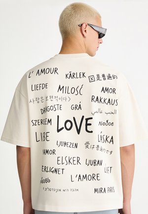 Persona con capelli biondo decolorati corti e occhiali da sole indossa una maglietta bianca con la parola "love" stampata sul retro in più lingue.