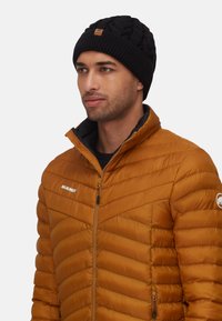 Beanie nera lavorata a maglia con motivo testurizzato e patch in pelle, abbinata a una giacca piumino ambrata trapuntata con tasche con zip.