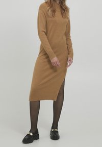 Vestido de manga larga color camel con cuello y puños acanalados, presenta una abertura lateral, combinado con mocasines negros y medias opacas.