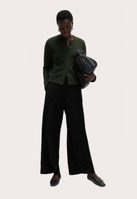 Mørkegrøn knap-op cardigan, sorte plisserede bukser med vide ben, sorte flade sko og en sort tekstureret taske, båret af en figur, der kigger ned.