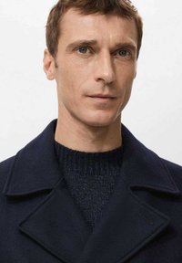 Abrigo de lana azul marino con diseño cruzado y solapas anchas, llevado sobre un suéter oscuro de punto. Textura suave y detalles mínimos.