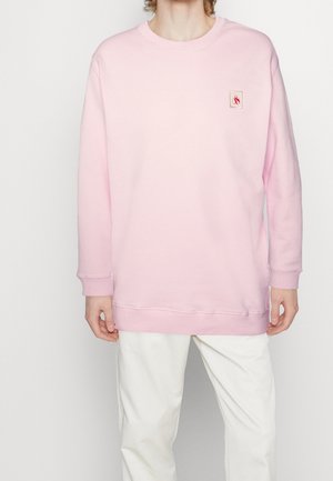 Personne portant un sweat-shirt oversize rose clair avec un petit patch logo rouge sur la poitrine gauche et un pantalon blanc, debout devant un fond uni.