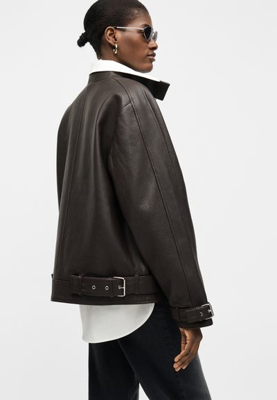 Γυναίκα με σκούρο καφέ δερμάτινο μπουφάν με αγκράφες, ορατό λευκό γιακά πουκαμίσου, μαύρο παντελόνι και γυαλιά ηλίου, κοιτάζοντας προφίλ σε απλό φόντο.