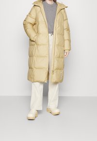 Person, der einen langen beigefarbenen Daunenmantel mit Kapuze über einem grauen Shirt, weißen Hosen und hellen Holzclogs tr�ägt, vor einem schlichten Hintergrund steht.