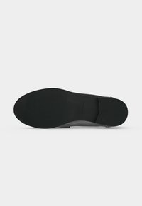 Semelle de chaussure noire avec une surface texturée au niveau des orteils et du talon, vue par dessous sur un fond clair uni.