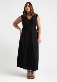 Abito maxi nero senza maniche con un design avvolgente, vita cinte e tessuto morbido; indossato con sandali con straps, presenta una silhouette liscia e fluida.