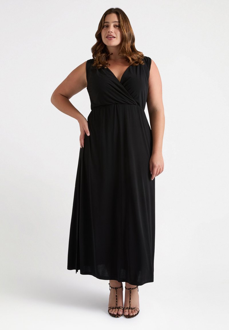 Abito maxi nero senza maniche con un design avvolgente, vita cinte e tessuto morbido; indossato con sandali con straps, presenta una silhouette liscia e fluida.