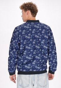 Marine bomberjack met een wit palmbomenpatroon, geribbelde zwarte kraag en manchetten, en een ontspannende pasvorm, gecombineerd met lichtdenim shorts.