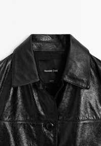 Zwart leren jack met een gladde, glanzende textuur, een kraag en een enkele knoopsluiting. Branding zichtbaar op het binnenlabel.