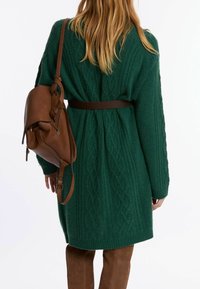 Groene kabelgebreide cardigan met een ceintuurlijn in de taille, gecombineerd met een bruine leren handtas. De stof lijkt zacht en met textuur, met een comfortabele pasvorm.