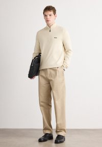 Light beige zip-neck sweater with black logo, paired with tan trousers and black leather shoes. Model holds a black bag.