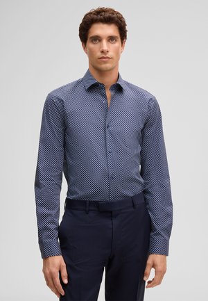 Mann trägt ein tailliertes, marineblau gemustertes Hemd mit Knöpfen und dunkelblaue Hosen, steht vor einem schlichten hellgrauen Hintergrund.