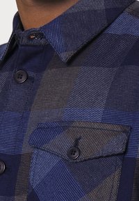 Koszula zapinana na guziki w niebieską kratkę. Wykonana z teksturowanej tkaniny, z gładkim kołnierzykiem i kieszonką na piersi, akcentowana ciemnymi guzikami.