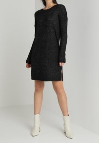 Zwarte gebreide jurk met een ontspannen pasvorm, lange mouwen en zijwaartse splitten. Gecombineerd met witte enkellaarsjes. Zachte textuur en losse silhouet.