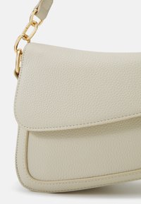 Helle beige Lederhandtasche mit strukturiertem Finish, Klappenverschluss und goldener Kettenriemen. Sie verfügt über abgerundete Kanten und sichtbare Nähte.