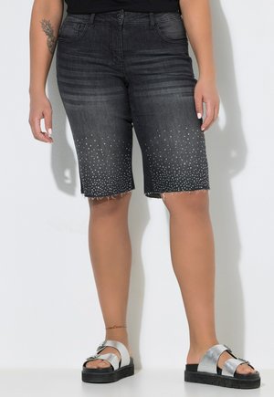 Person trägt dunkelgraue, knielange Denim-Shorts mit weißen Punkt-Details, schwarzes Top, silberne Plateau-Sandalen und ein Fußkettchen.