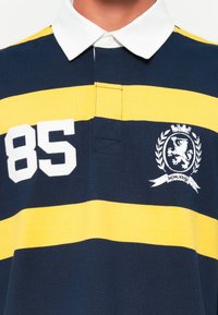 Tamsiai mėlynas ir geltonas juostuotas regbio marškinėliai su balta apykakle, kuriuose pavaizduotas numeris "85" ir liūto emblema su laurų vainiku.