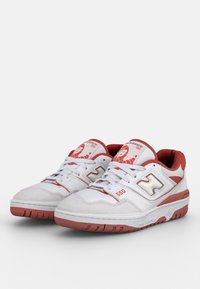 Baskets blanches avec des accents rouges, présentant une tige en cuir perforé, un col rembourré et une semelle en caoutchouc. Le numéro de modèle "550" est visible sur le côté.