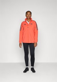 Heldere oranje jas met hoge kraag, ritssluiting en marineblauwe strepen op de mouwen. Draagt een zwarte legging en sportschoenen.