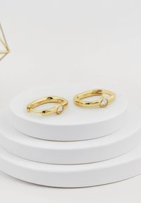 Guldaktiga hoopörhängen med en slät yta, som har en ensam klar sten i en rund design. Visas på vita cirkulära ställ.