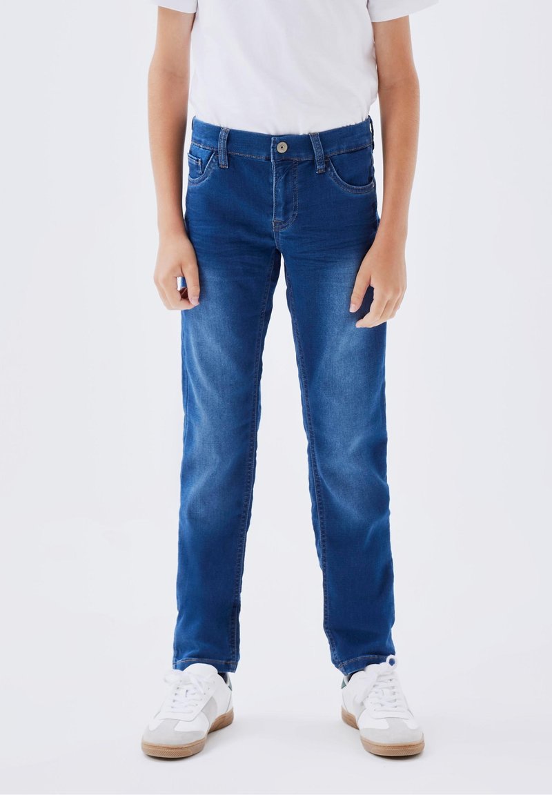 Enfant portant un jean bleu, des baskets blanches et un t-shirt blanc, debout devant un fond blanc uni.
