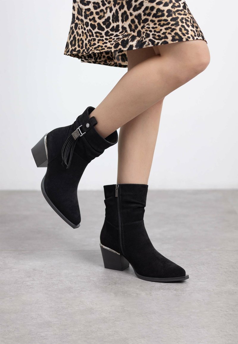 Botines de ante negros con un acabado texturizado, un zipper lateral y un detalle plateado en el tacón, combinados con una falda de estampado de leopardo.