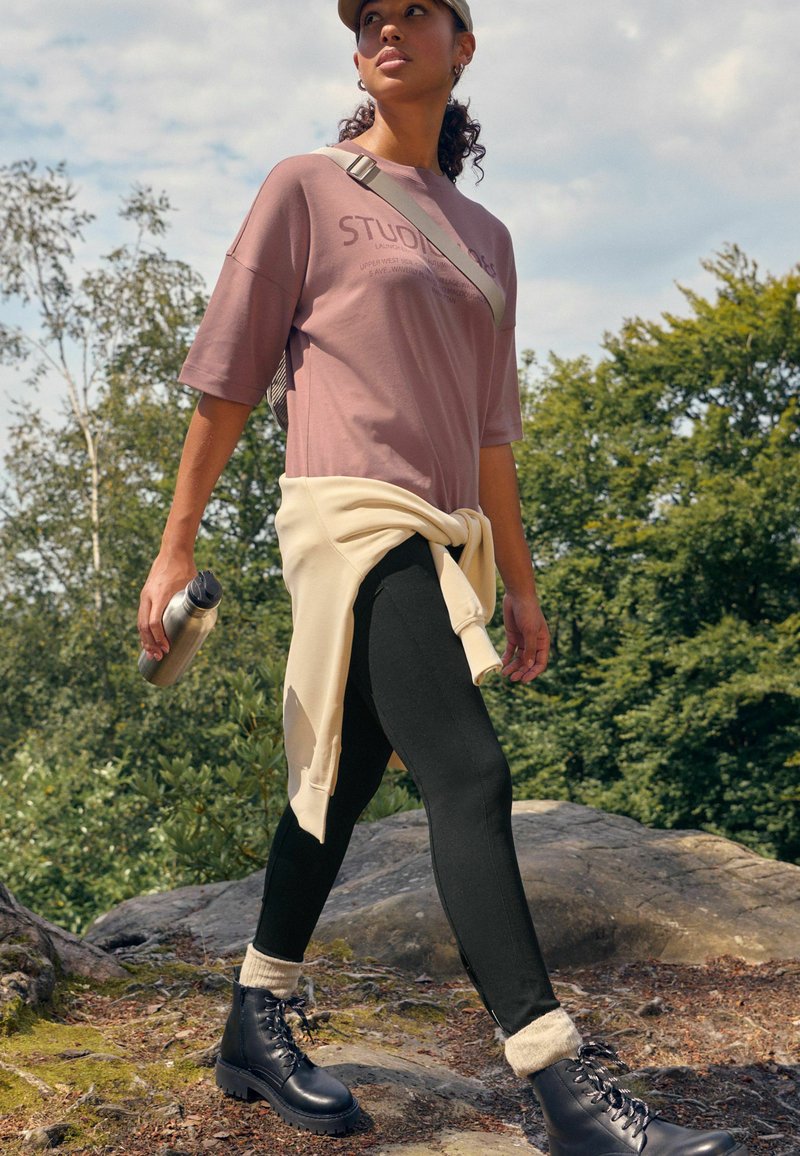 T-shirt rose oversize avec texte, leggings noirs, sweatshirt crème noué à la taille, bottes noires à lacets, et une bouteille d'eau, à l'extérieur.
