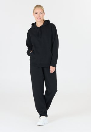 Schwarzer Hoodie mit einer Fronttasche und Kordeln, kombiniert mit lockeren schwarzen Jogginghosen. Das Model trägt weiße Sneaker. Glatte Textur.