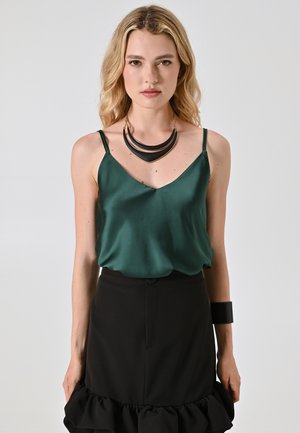 Grüne Satinkamer mit dünnen Trägern, kombiniert mit einem schwarzen Rüschensrock. Als Accessoires dienen ein klobiges schwarzes Armband und eine mehrlagige Halskette.