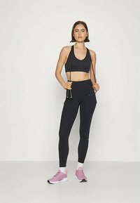 Kvinne i svart sports-bh og tights holder et svart hoppetau, iført hvite sokker og rosa joggesko, stående mot en ensfarget bakgrunn.