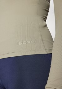 Hosszú ujjú felső világos olívazöld bordás anyagból, logóval az alján. Sötét tengerészkék, magas derekú leggingsszel párosítva.