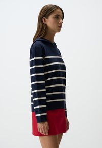 Felpa con cappuccio blu marina con strisce bianche, realizzata in tessuto morbido. Abbinata a una mini gonna rossa. Vista laterale, la modella è in piedi con espressione neutra.