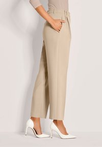 Vrouw in beige hoog-wijdte op maat gemaakte broek en witte puntige hoge hakken, staande met één hand in de zak.