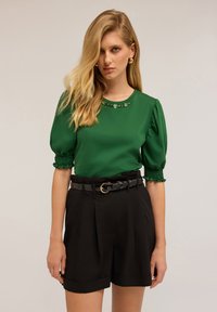 Top verde de manga corta con detalles fruncidos, escote redondo y ribete adornado. Combinado con pantalones cortos negros de tiro alto y un cinturón negro.
