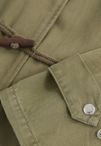 Koszula z zielonej oliwkowej bawełny z kołnierzykiem w formie stójki, zapięciem na guziki oraz drewnianym zapięciem w formie toggle z plecionym sznurkiem, prezentująca teksturowaną tkaninę.
