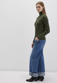 Dunkelgrüner, kabelgestrickter Rollkragenpullover kombiniert mit lockeren blauen Jeans mit umgeschlagenen Säumen und schwarzen hohen Stiefeln.