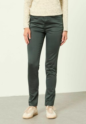 Personne portant un pantalon slim vert foncé, un haut beige à manches longues et des baskets beige clair, debout sur un sol gris.