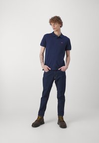 Marineblå polo skjorte med krage og korte ermer, kombinert med mørke denimjeans og brune snørestøvler med gule snører.