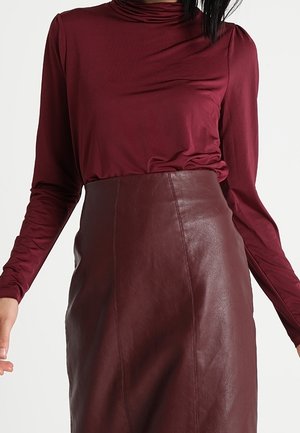 Femme portant un haut bordeaux à manches longues rentré dans une jupe en cuir marron foncé taille haute sur un fond uni.