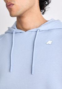 Gros plan d'une personne portant un sweat à capuche bleu clair avec des cordons et un petit logo blanc sur la poitrine.