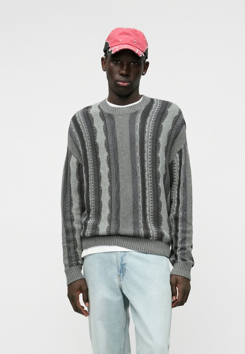 Γκρίζος πουλόβερ με κάθετες κυματιστές ρίγες, με ριμπ φόδρα στους καρπούς και το τελείωμα, φορεμένο πάνω από λευκό t-shirt; συνδυασμένο με ανοιχτό γαλάζιο τζιν και ροζ καπέλο.