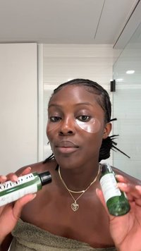 Deux flacons de soins de la peau verts sont tenus par une personne à la peau foncée et aux tresses. La personne porte des patchs sous les yeux et une serviette. Fond neutre de salle de bain.