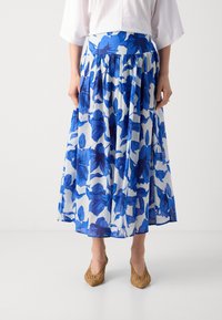 Jupe florale bleue avec un fond blanc, longueur genoux, avec une taille froncée et un tissu léger, associée à des talons tissés beiges.
